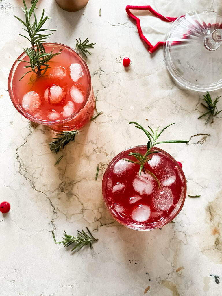 santa's tipsy spritzer cocktail in a whisky glass with rosemary as garnish, a measuring cup and cranberries in a glass coupe in the background