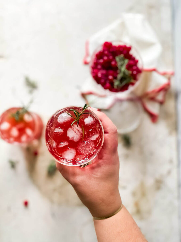 santa's tipsy spritzer cocktail in a whisky glass with rosemary as garnish, a measuring cup and cranberries in a glass coupe in the background