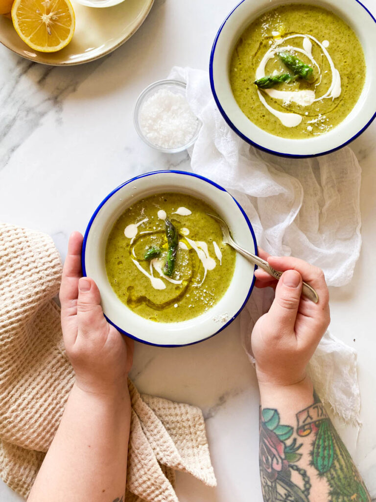 Broccoli Asparagus Soup: Delicious Lunch Idea 1 on a marble background, two towels and two bowl of broccoli asparagus soup, two hands holding one of the bowls