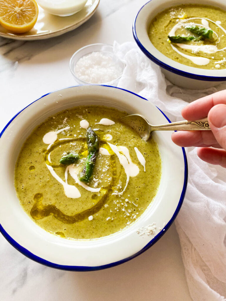 Broccoli Asparagus Soup: Delicious Lunch Idea 3 lemon, salt & cream in a bowl, on a marble background, two towels and two bowl of broccoli asparagus soup, hand holding a spoon