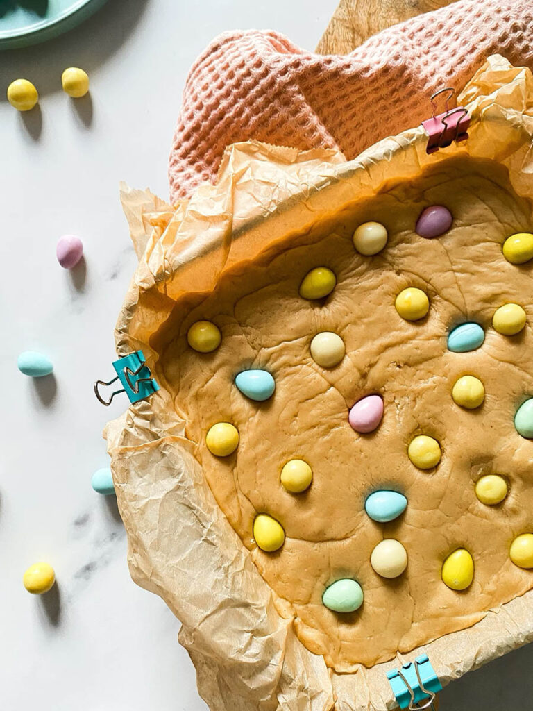 Easter Mini Eggs Fudge, Delicious And With No Condensed Milk 1 Mini eggs fudge in a tray over a pink napkin and with mini chocolate eggs scattered around the marble countertop