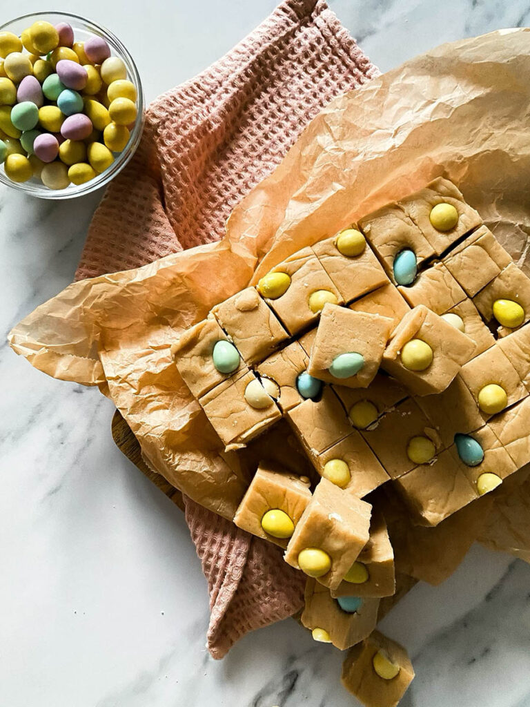Easter Mini Eggs Fudge, Delicious And With No Condensed Milk 4 Mini eggs fudge chunks on parchment paper over a pink napkin and with mini chocolate eggs scattered around the marble countertop