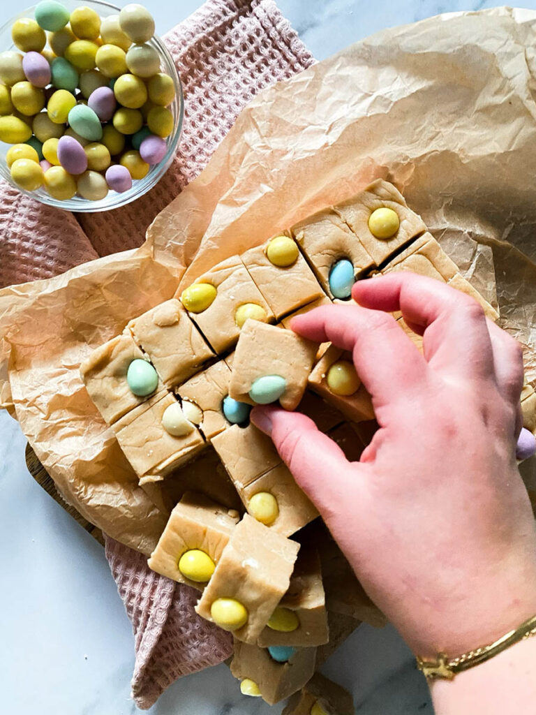 Easter Mini Eggs Fudge, Delicious And With No Condensed Milk 3 Mini eggs fudge chunks on parchment paper over a pink napkin and with mini chocolate eggs scattered around the marble countertop