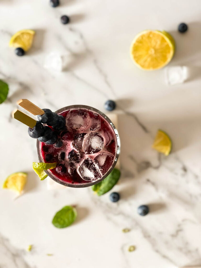 Delicious Blueberry Mojito Mocktail Recipe 4 blueberry mojito mocktail glass from above on a marble square drink holder with fresh mint, ice and fresh lime scattered around