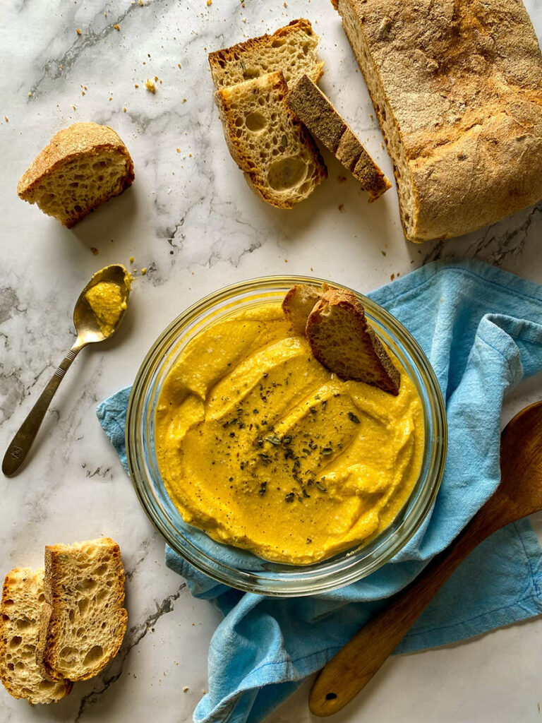 5 Ingredients Pumpkin Whipped Feta Dip, Delicious Fall Dip 1 A bowl of creamy pumpkin whipped feta dip topped with herbs, surrounded by slices of crusty bread on a marble surface. A spoon with some of the dip rests nearby, and a loaf of rustic bread is placed on the side. The setting includes a light blue napkin and a wooden spoon for a casual, rustic presentation.