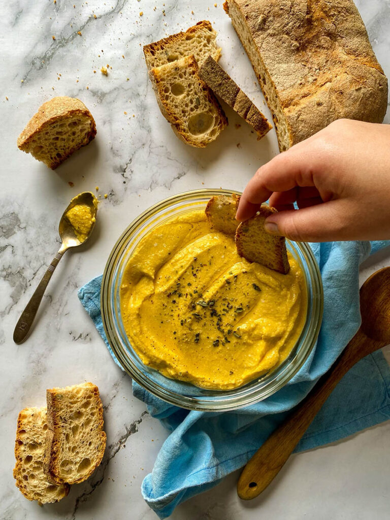5 Ingredients Pumpkin Whipped Feta Dip, Delicious Fall Dip 2 A bowl of creamy pumpkin whipped feta dip topped with herbs, surrounded by slices of crusty bread on a marble surface. A spoon with some of the dip rests nearby, and a loaf of rustic bread is placed on the side. The setting includes a light blue napkin and a wooden spoon for a casual, rustic presentation.