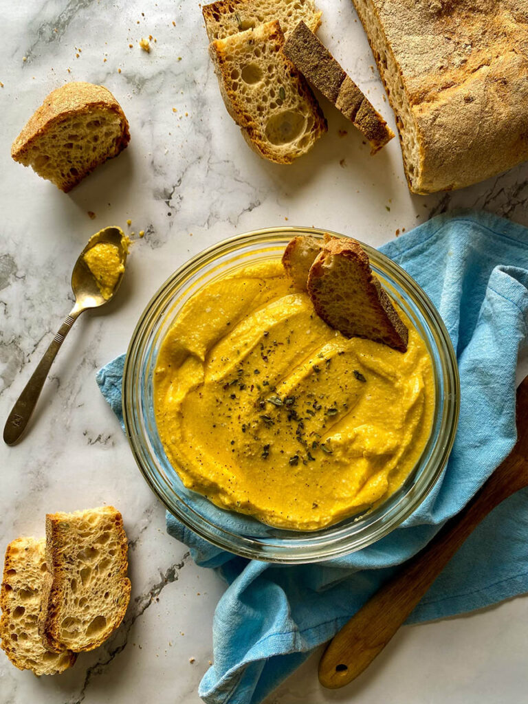 5 Ingredients Pumpkin Whipped Feta Dip, Delicious Fall Dip 3 A bowl of creamy pumpkin whipped feta dip topped with herbs, surrounded by slices of crusty bread on a marble surface. A spoon with some of the dip rests nearby, and a loaf of rustic bread is placed on the side. The setting includes a light blue napkin and a wooden spoon for a casual, rustic presentation.