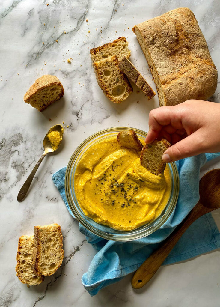 5 Ingredients Pumpkin Whipped Feta Dip, Delicious Fall Dip 5 A bowl of creamy pumpkin whipped feta dip topped with herbs, surrounded by slices of crusty bread on a marble surface. A spoon with some of the dip rests nearby, and a loaf of rustic bread is placed on the side. The setting includes a light blue napkin and a wooden spoon for a casual, rustic presentation.