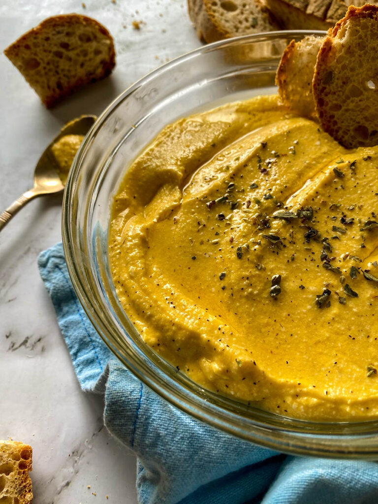 5 Ingredients Pumpkin Whipped Feta Dip, Delicious Fall Dip 6 A bowl of creamy pumpkin whipped feta dip topped with herbs, surrounded by slices of crusty bread on a marble surface. A spoon with some of the dip rests nearby, and a loaf of rustic bread is placed on the side. The setting includes a light blue napkin and a wooden spoon for a casual, rustic presentation.