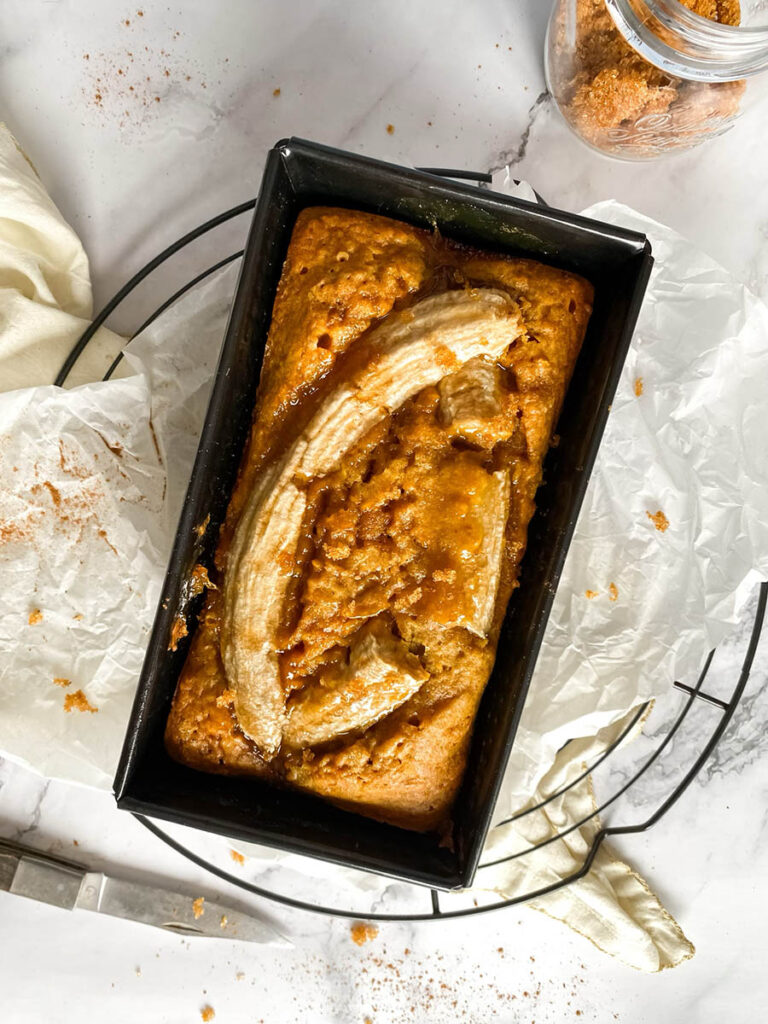 Vegan Pumpkin Banana Bread, Delicious And Easy 1 A loaf of freshly baked pumpkin banana bread in a black rectangular pan, topped with caramelized banana slices. The bread is surrounded by parchment paper, with a knife and scattered crumbs visible on the marble surface.