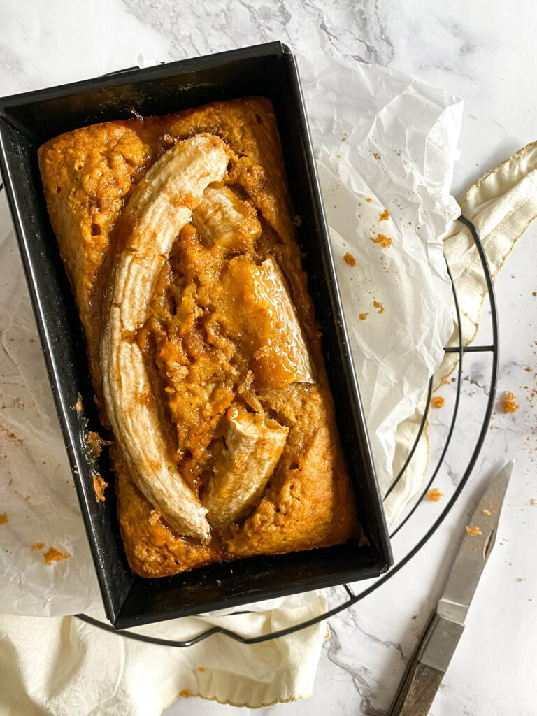 Vegan Pumpkin Banana Bread, Delicious And Easy 2 A loaf of freshly baked pumpkin banana bread in a black rectangular pan, topped with caramelized banana slices. The bread is surrounded by parchment paper, with a knife and scattered crumbs visible on the marble surface.