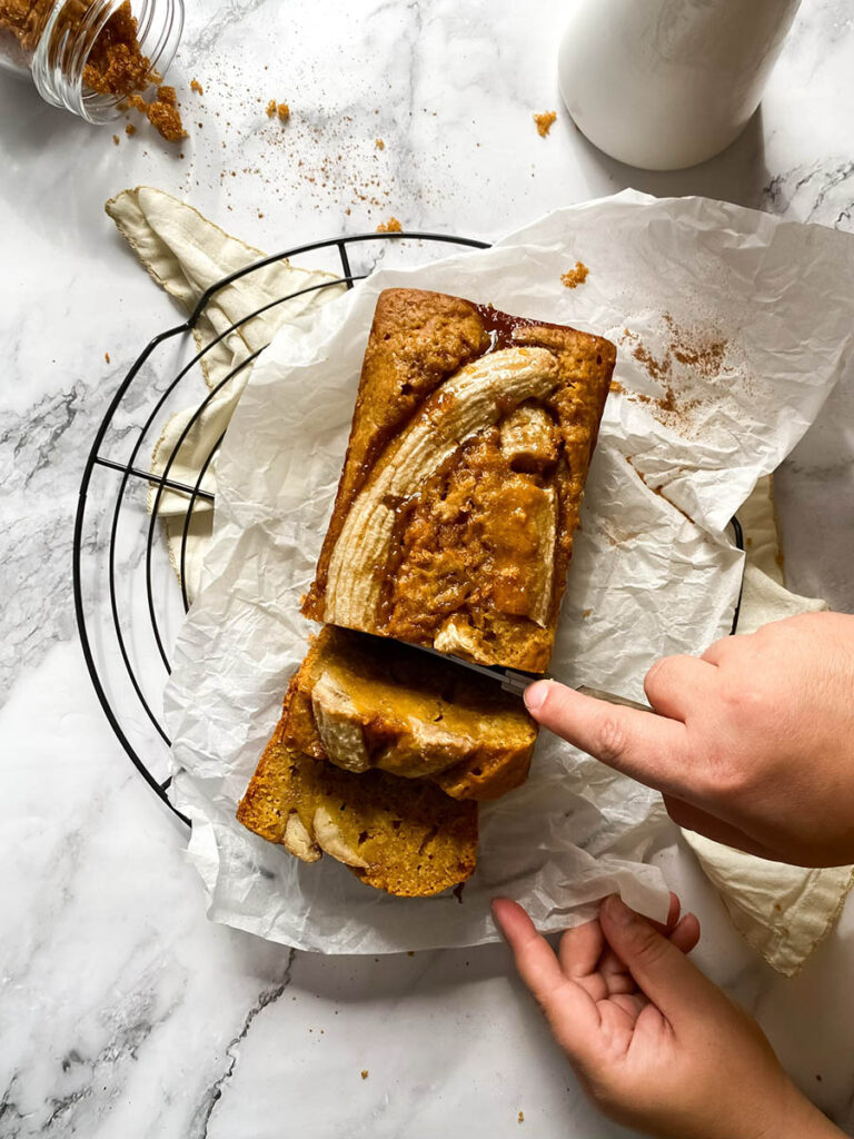 Vegan Pumpkin Banana Bread, Delicious And Easy 7 A loaf of pumpkin banana bread sliced on parchment paper, with a golden crust and caramelized banana on top. A jar of brown sugar is tipped over in the background, adding texture to the countertop.