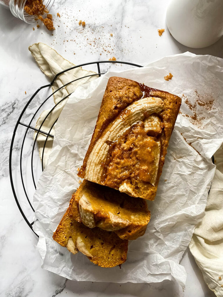 Vegan Pumpkin Banana Bread, Delicious And Easy 8 A loaf of pumpkin banana bread sliced on parchment paper, with a golden crust and caramelized banana on top. A jar of brown sugar is tipped over in the background, adding texture to the countertop.