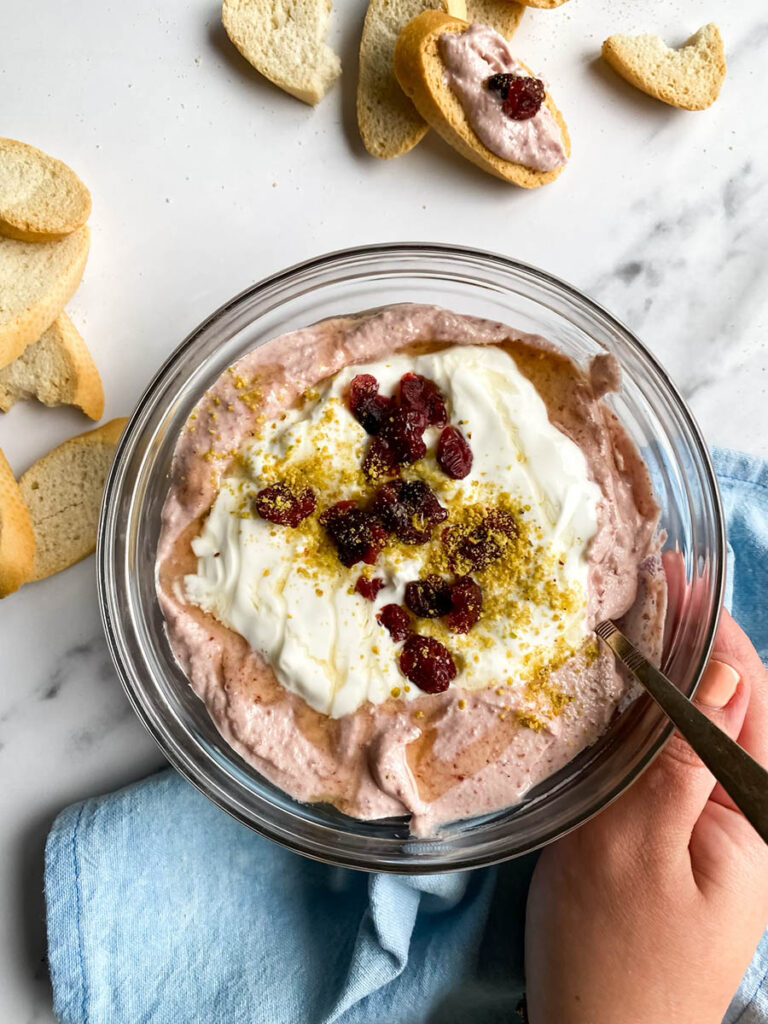 A glass bowl filled with cranberry whipped feta dip, topped with crushed pistachios, dried cranberries, and a drizzle of olive oil. Surrounding the bowl are crisp baguette slices and a small dish of whole pistachios, all set on a marble surface with a light blue cloth beside the bowl.