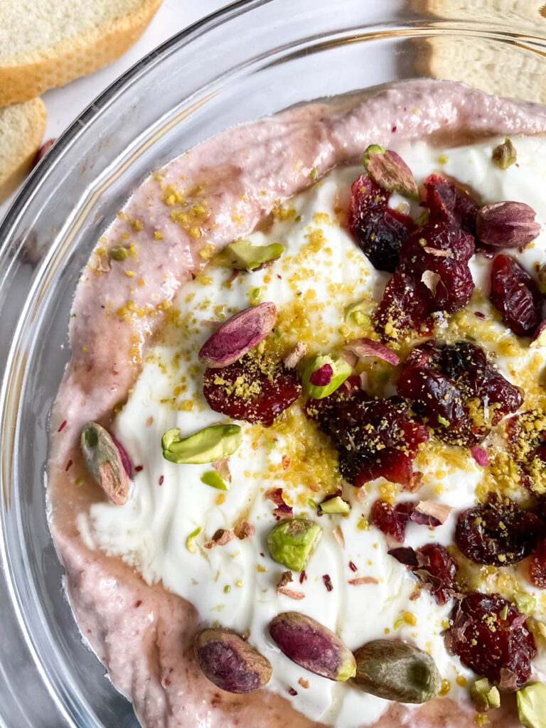 A glass bowl filled with cranberry whipped feta dip, topped with crushed pistachios, dried cranberries, and a drizzle of olive oil. Surrounding the bowl are crisp baguette slices and a small dish of whole pistachios, all set on a marble surface with a light blue cloth beside the bowl.