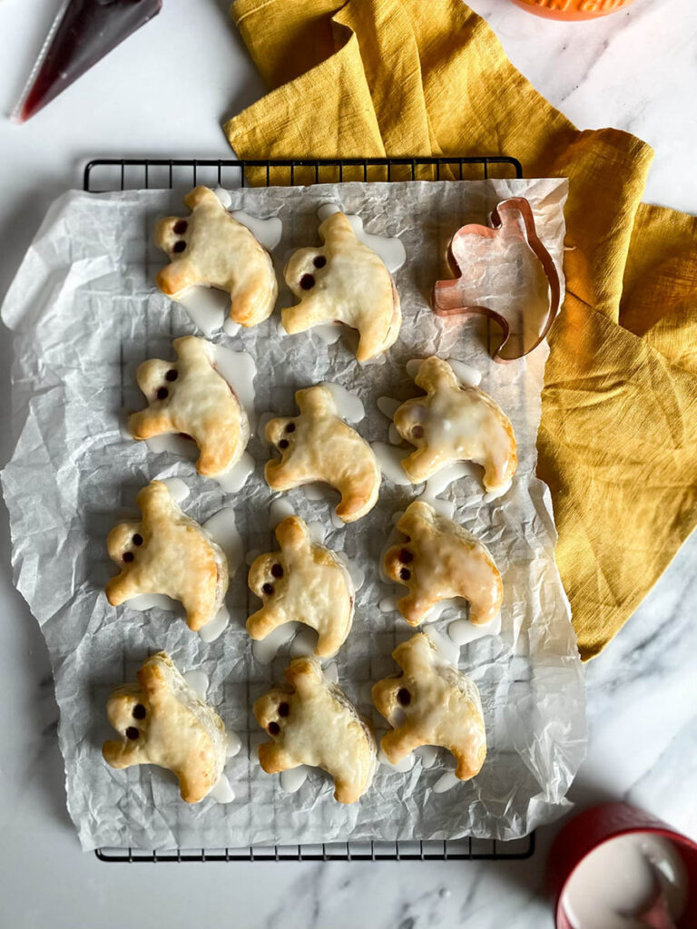 Ghost Hand Pies: Cute & Delicious Halloween Dessert 1 A tray of ghost hand pies is cooling on a wire rack covered with parchment paper. The pies are iced with a white glaze, and their "eyes" are made with small dark filling dots. A copper ghost-shaped cookie cutter and a yellow kitchen towel are placed nearby for decoration.