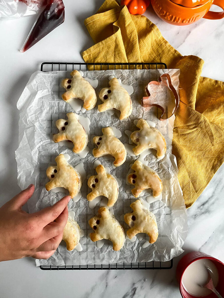 Ghost Hand Pies: Cute & Delicious Halloween Dessert 4 A tray of ghost hand pies is cooling on a wire rack covered with parchment paper. The pies are iced with a white glaze, and their "eyes" are made with small dark filling dots. A copper ghost-shaped cookie cutter and a yellow kitchen towel are placed nearby for decoration.
