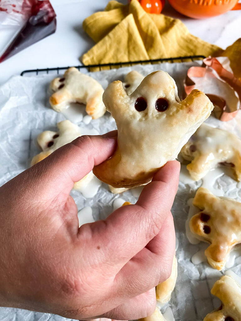 Ghost Hand Pies: Cute & Delicious Halloween Dessert 3 A hand holds a single ghost-shaped hand pie with white glaze and two small dark filling dots for eyes. The background shows more ghost pies cooling on a tray, along with a copper ghost cookie cutter and a yellow cloth.