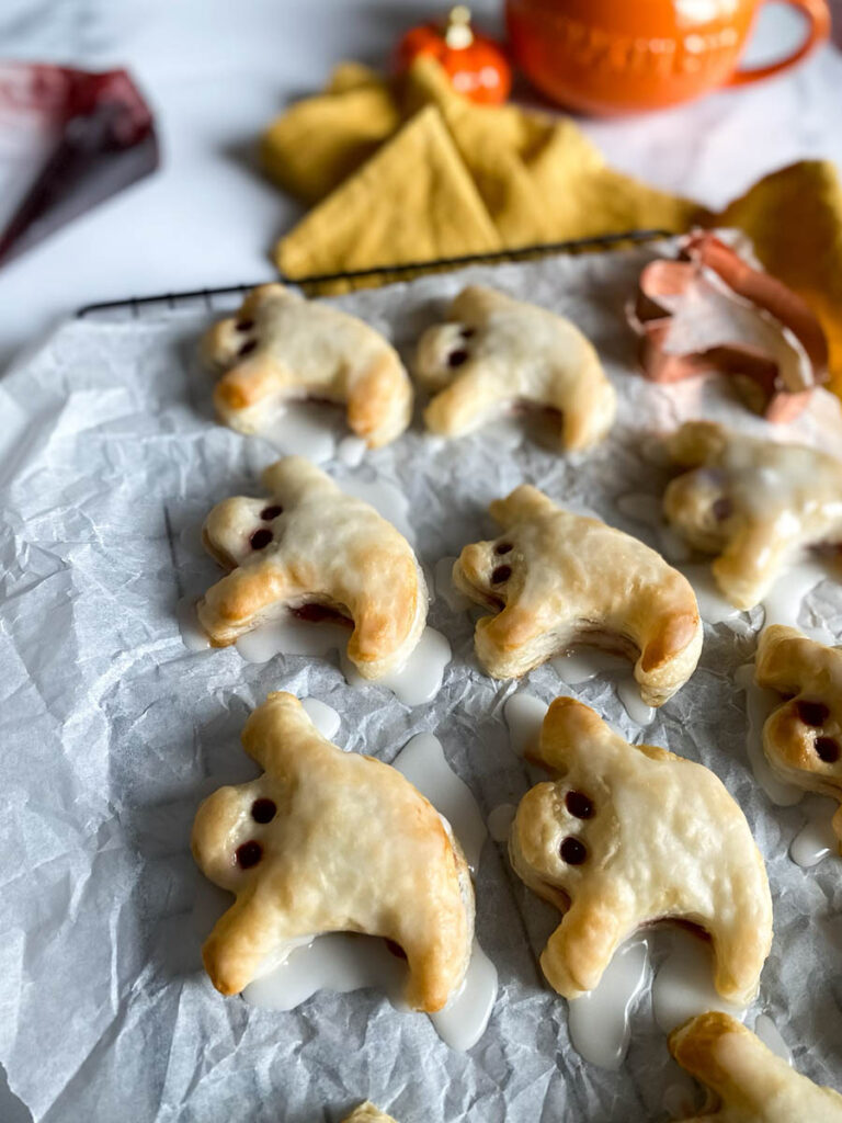 Ghost Hand Pies: Cute & Delicious Halloween Dessert 8 A tray of ghost hand pies is cooling on a wire rack covered with parchment paper. The pies are iced with a white glaze, and their "eyes" are made with small dark filling dots. A copper ghost-shaped cookie cutter and a yellow kitchen towel are placed nearby for decoration.