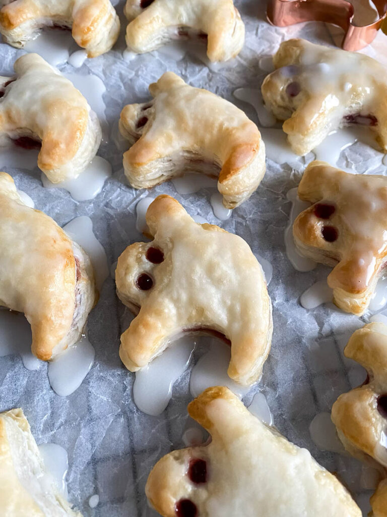 Ghost Hand Pies: Cute & Delicious Halloween Dessert 9 A tray of ghost hand pies is cooling on a wire rack covered with parchment paper. The pies are iced with a white glaze, and their "eyes" are made with small dark filling dots. A copper ghost-shaped cookie cutter and a yellow kitchen towel are placed nearby for decoration.