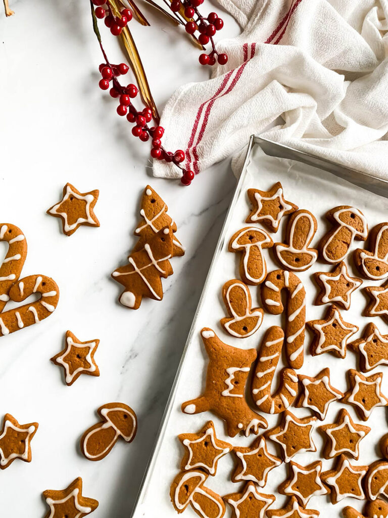 Adorable Christmas Gingerbread Ornaments, Easy & Quick 1 A tray of homemade. Christmas gingerbread ornaments decorated with white icing in festive shapes like stars, trees, mushrooms, and candy canes. Some cookies are scattered outside the tray on a white countertop, with red berry stems and a white cloth with red stripes in the background, giving a cozy holiday vibe.