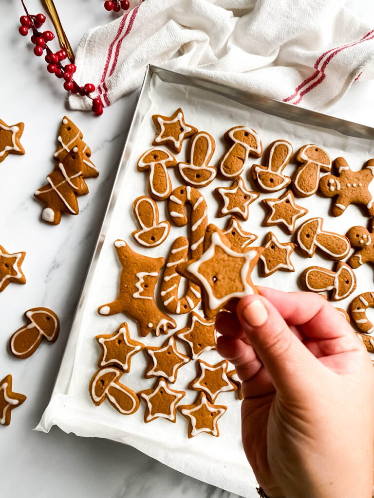 Adorable Christmas Gingerbread Ornaments, Easy & Quick 7 A tray of homemade. Christmas gingerbread ornaments decorated with white icing in festive shapes like stars, trees, mushrooms, and candy canes. Some cookies are scattered outside the tray on a white countertop, with red berry stems and a white cloth with red stripes in the background, giving a cozy holiday vibe.