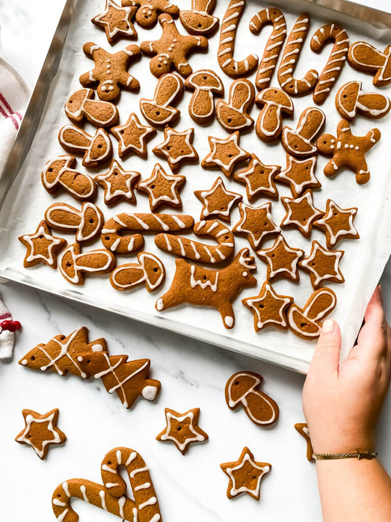 Adorable Christmas Gingerbread Ornaments, Easy & Quick 8 A tray of homemade. Christmas gingerbread ornaments decorated with white icing in festive shapes like stars, trees, mushrooms, and candy canes. Some cookies are scattered outside the tray on a white countertop, with red berry stems and a white cloth with red stripes in the background, giving a cozy holiday vibe.