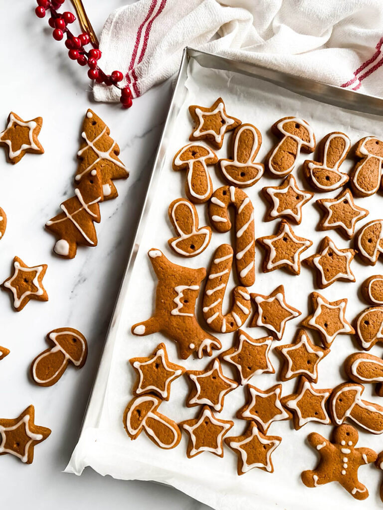 Adorable Christmas Gingerbread Ornaments, Easy & Quick 4 A tray of homemade. Christmas gingerbread ornaments decorated with white icing in festive shapes like stars, trees, mushrooms, and candy canes. Some cookies are scattered outside the tray on a white countertop, with red berry stems and a white cloth with red stripes in the background, giving a cozy holiday vibe.