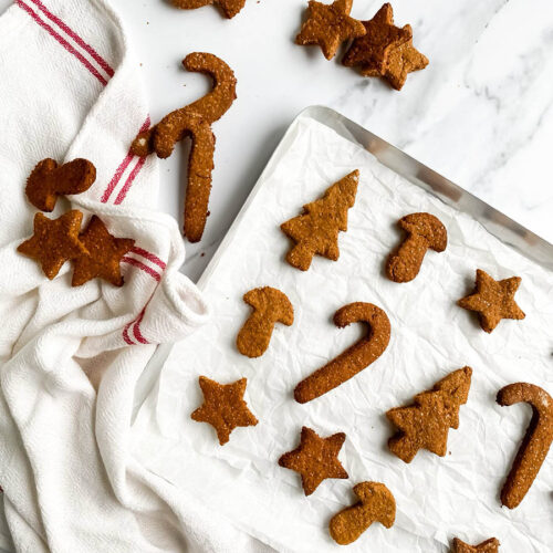 Homemade gingerbread cookies for dogs shaped like stars, candy canes, and Christmas trees, arranged on parchment paper and a white towel with red stripes. These rustic, wholesome treats make a festive, dog-friendly snack option for the holiday season.