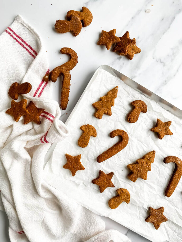 Homemade gingerbread cookies for dogs shaped like stars, candy canes, and Christmas trees, arranged on parchment paper and a white towel with red stripes. These rustic, wholesome treats make a festive, dog-friendly snack option for the holiday season.