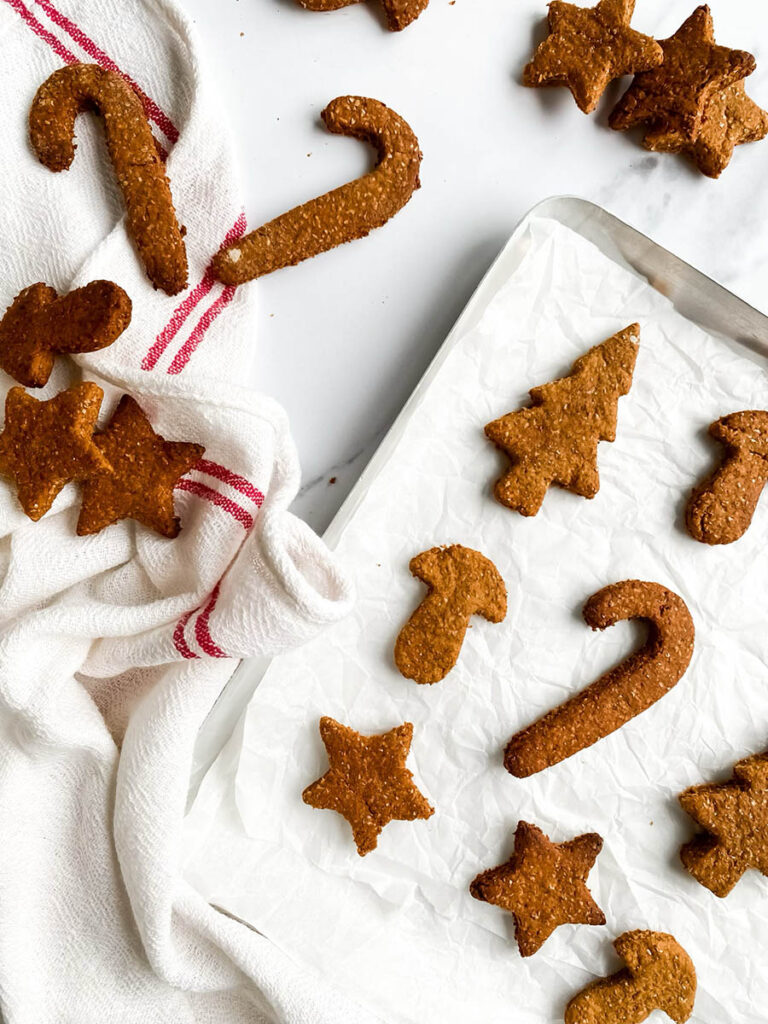 Homemade gingerbread cookies for dogs shaped like stars, candy canes, and Christmas trees, arranged on parchment paper and a white towel with red stripes. These rustic, wholesome treats make a festive, dog-friendly snack option for the holiday season.