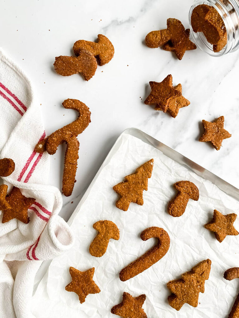 Homemade gingerbread cookies for dogs shaped like stars, candy canes, and Christmas trees, arranged on parchment paper and a white towel with red stripes. These rustic, wholesome treats make a festive, dog-friendly snack option for the holiday season.