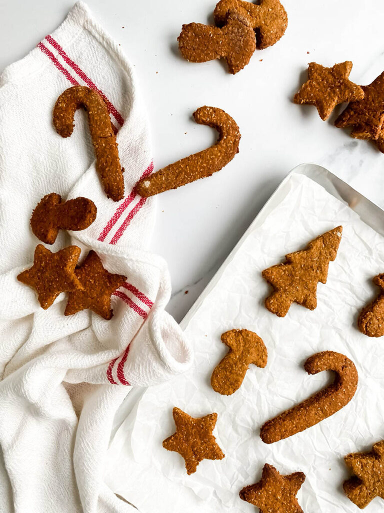 Homemade gingerbread cookies for dogs shaped like stars, candy canes, and Christmas trees, arranged on parchment paper and a white towel with red stripes. These rustic, wholesome treats make a festive, dog-friendly snack option for the holiday season.