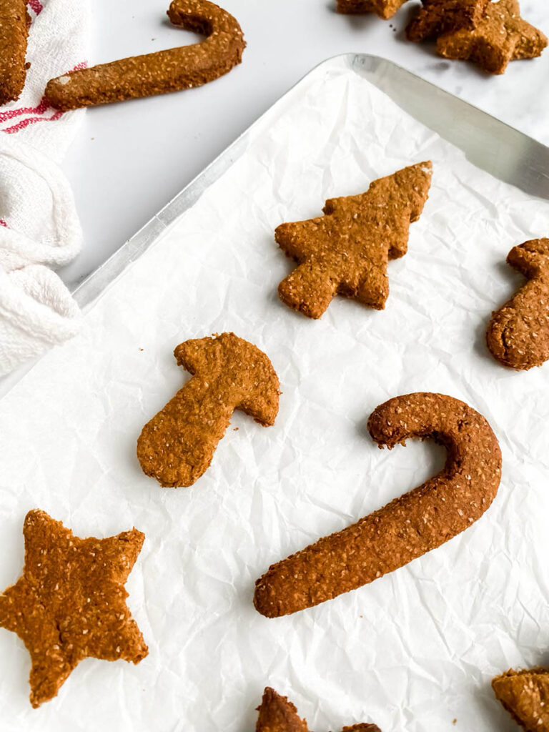 Homemade gingerbread cookies for dogs shaped like stars, candy canes, and Christmas trees, arranged on parchment paper and a white towel with red stripes. These rustic, wholesome treats make a festive, dog-friendly snack option for the holiday season.
