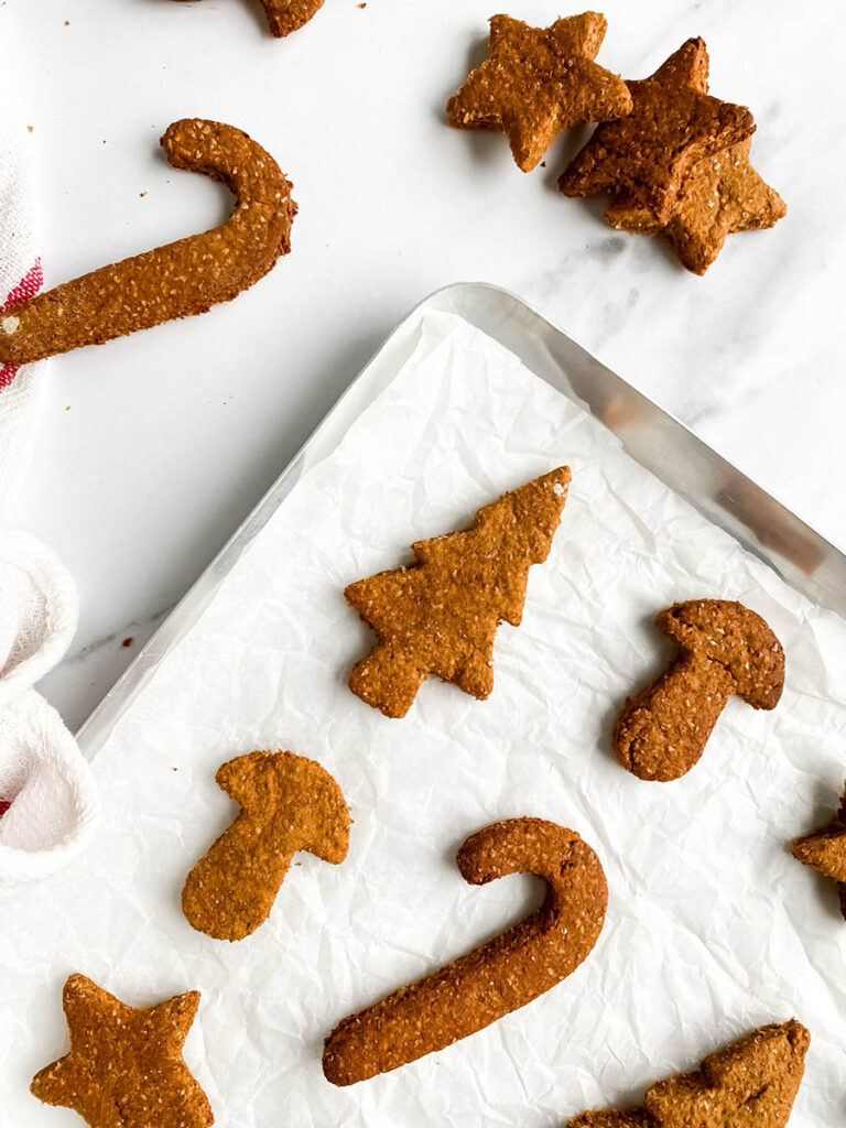 Homemade gingerbread cookies for dogs shaped like stars, candy canes, and Christmas trees, arranged on parchment paper and a white towel with red stripes. These rustic, wholesome treats make a festive, dog-friendly snack option for the holiday season.