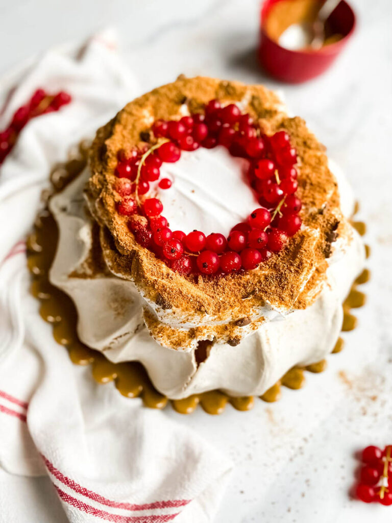 Gingerbread Pavlova, Delicious Christmas Dessert 1 A beautifully styled gingerbread pavlova topped with a wreath of vibrant red currants sits on a white surface. The dessert features a crisp, meringue-like base with a dusting of gingerbread crumbs, creating a rustic yet elegant presentation. A red and white striped cloth adds a festive touch to the composition.