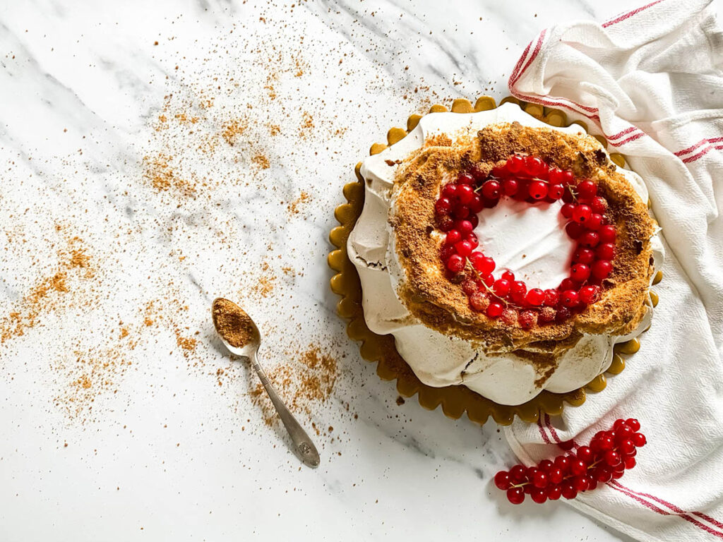 Gingerbread Pavlova, Delicious Christmas Dessert 4 A beautifully styled gingerbread pavlova topped with a wreath of vibrant red currants sits on a white surface. The dessert features a crisp, meringue-like base with a dusting of gingerbread crumbs, creating a rustic yet elegant presentation. A red and white striped cloth adds a festive touch to the composition.