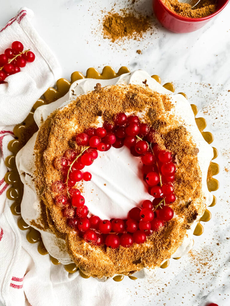 Gingerbread Pavlova, Delicious Christmas Dessert 3 A beautifully styled gingerbread pavlova topped with a wreath of vibrant red currants sits on a white surface. The dessert features a crisp, meringue-like base with a dusting of gingerbread crumbs, creating a rustic yet elegant presentation. A red and white striped cloth adds a festive touch to the composition.