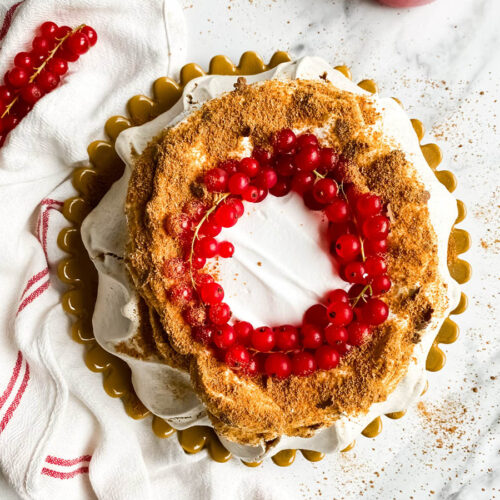 Gingerbread Pavlova, Delicious Christmas Dessert 6 from top - A beautifully styled gingerbread pavlova topped with a wreath of vibrant red currants sits on a white surface. The dessert features a crisp, meringue-like base with a dusting of gingerbread crumbs, creating a rustic yet elegant presentation. A red and white striped cloth adds a festive touch to the composition.
