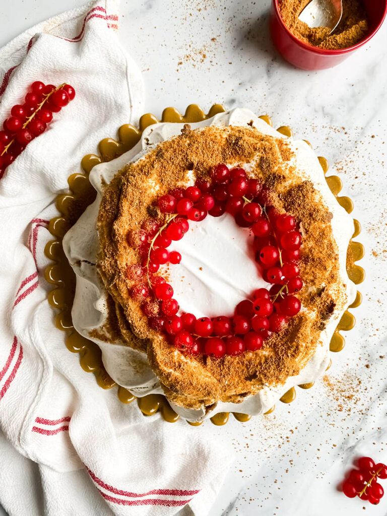 Gingerbread Pavlova, Delicious Christmas Dessert 5 A beautifully styled gingerbread pavlova topped with a wreath of vibrant red currants sits on a white surface. The dessert features a crisp, meringue-like base with a dusting of gingerbread crumbs, creating a rustic yet elegant presentation. A red and white striped cloth adds a festive touch to the composition.