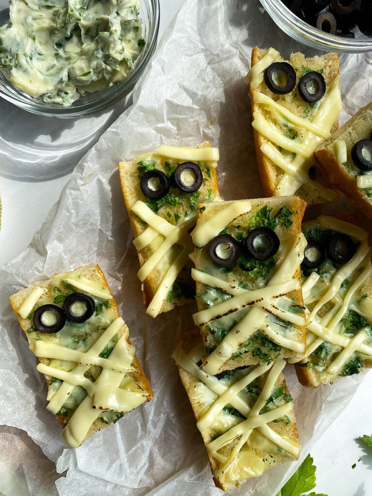 Mummy Garlic Bread, Deliciously Scary Halloween Snack 2 An overhead view of mummy-themed garlic bread slices topped with melted cheese and black olive slices arranged to resemble eyes. The bread sits on parchment paper, with bowls of black olive slices and a creamy herb spread beside it. Fresh parsley, garlic cloves, and a cloth napkin are placed around the arrangement for decoration.