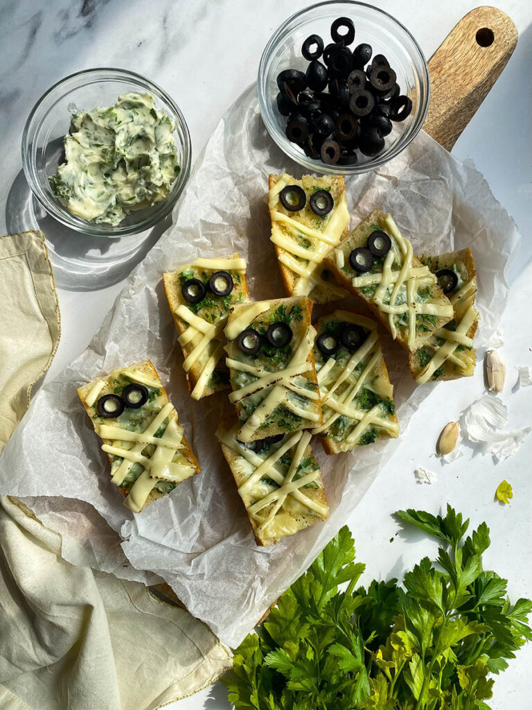Mummy Garlic Bread, Deliciously Scary Halloween Snack 3 An overhead view of mummy-themed garlic bread slices topped with melted cheese and black olive slices arranged to resemble eyes. The bread sits on parchment paper, with bowls of black olive slices and a creamy herb spread beside it. Fresh parsley, garlic cloves, and a cloth napkin are placed around the arrangement for decoration.
