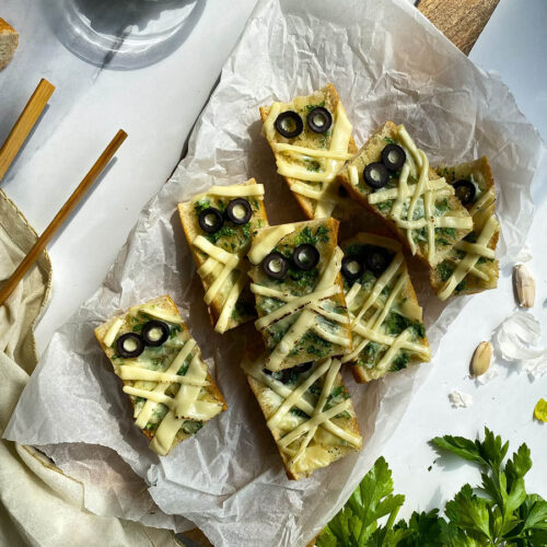 Mummy Garlic Bread, Deliciously Scary Halloween Snack 4 mummy garlic bread pieces on a wooden board, parsley around, a bowl with garlic butter and a bowl with olives in the back, towel and garlic around