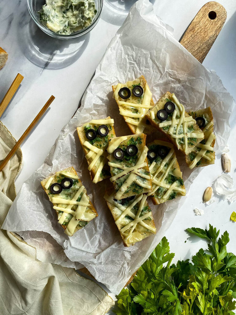 Mummy Garlic Bread, Deliciously Scary Halloween Snack 1 An overhead view of mummy-themed garlic bread slices topped with melted cheese and black olive slices arranged to resemble eyes. The bread sits on parchment paper, with bowls of black olive slices and a creamy herb spread beside it. Fresh parsley, garlic cloves, and a cloth napkin are placed around the arrangement for decoration.