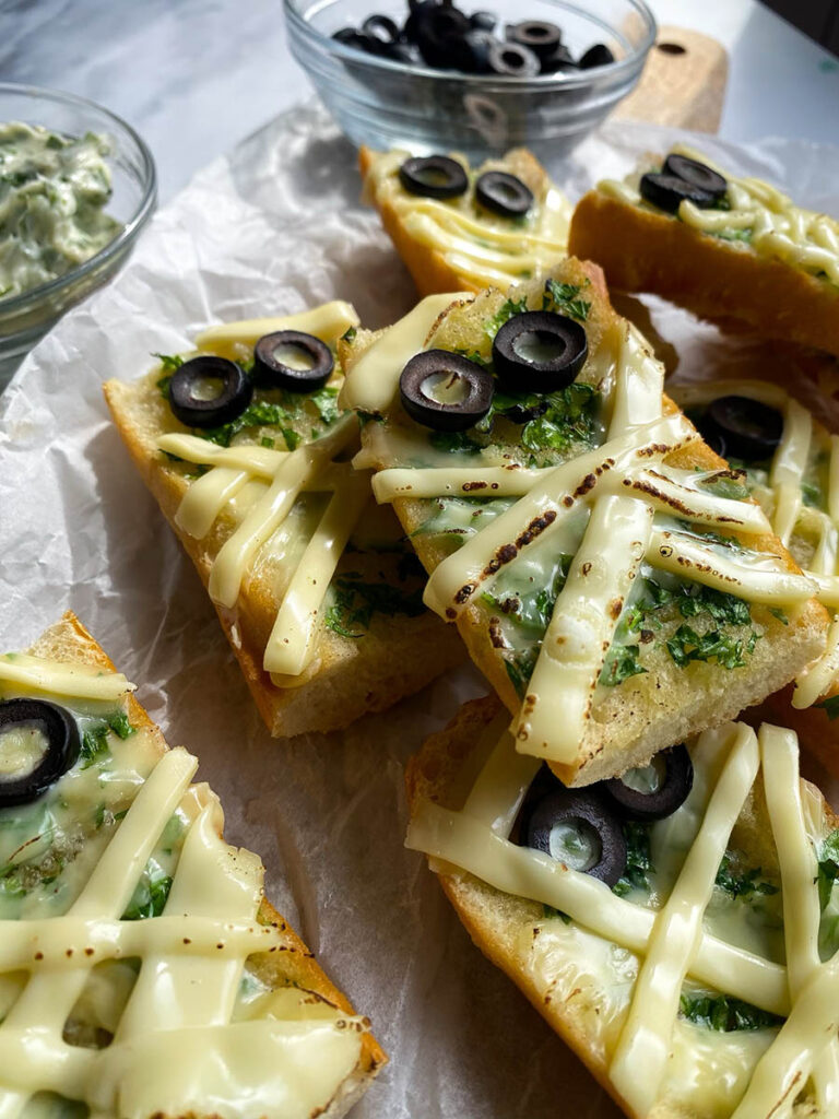 Mummy Garlic Bread, Deliciously Scary Halloween Snack 6 An overhead view of mummy-themed garlic bread slices topped with melted cheese and black olive slices arranged to resemble eyes. The bread sits on parchment paper, with bowls of black olive slices and a creamy herb spread beside it. Fresh parsley, garlic cloves, and a cloth napkin are placed around the arrangement for decoration.