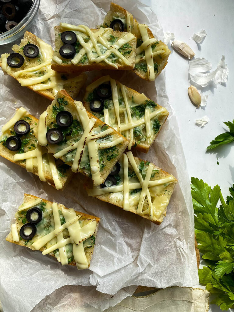 Mummy Garlic Bread, Deliciously Scary Halloween Snack 7 An overhead view of mummy-themed garlic bread slices topped with melted cheese and black olive slices arranged to resemble eyes. The bread sits on parchment paper, with bowls of black olive slices and a creamy herb spread beside it. Fresh parsley, garlic cloves, and a cloth napkin are placed around the arrangement for decoration.