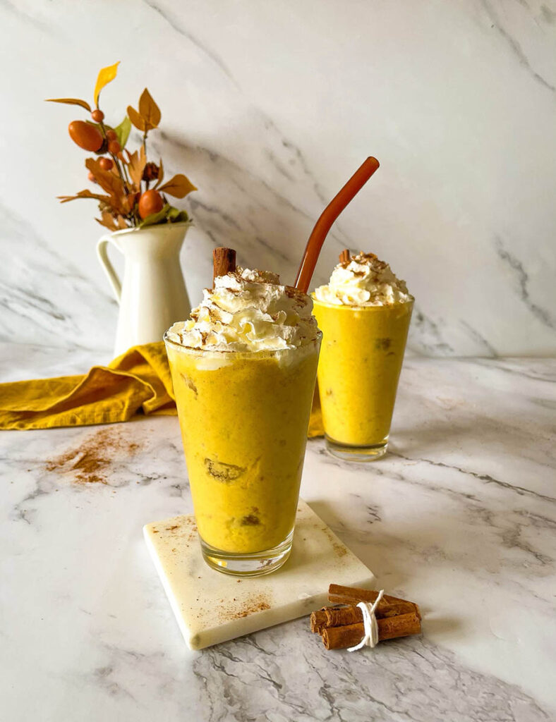 Irresistible Pumpkin Spice Mocktail Recipe 7 Two tall glasses of creamy pumpkin spice mocktail topped with whipped cream and garnished with cinnamon sticks, set on a marble surface. A bundle of cinnamon sticks and a yellow cloth add cozy fall decor in the background, along with a small vase of autumn leaves.