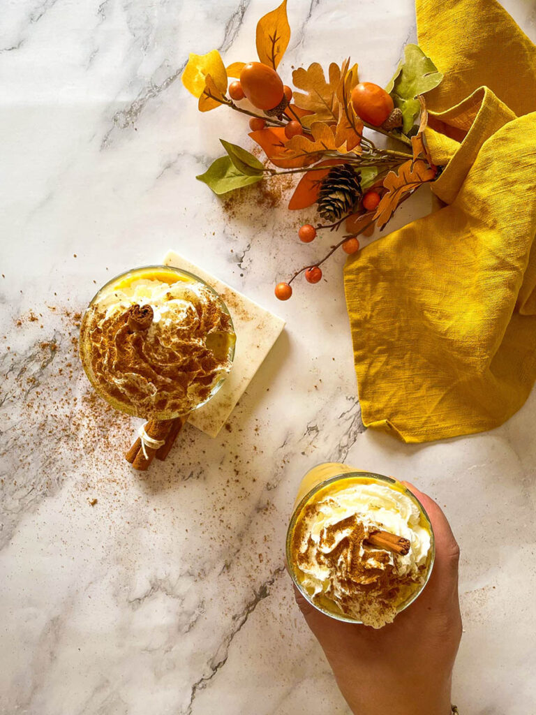 Irresistible Pumpkin Spice Mocktail Recipe 3 Two tall glasses of creamy pumpkin spice mocktail topped with whipped cream and garnished with cinnamon sticks, set on a marble surface. A bundle of cinnamon sticks and a yellow cloth add cozy fall decor in the background, along with a small vase of autumn leaves.