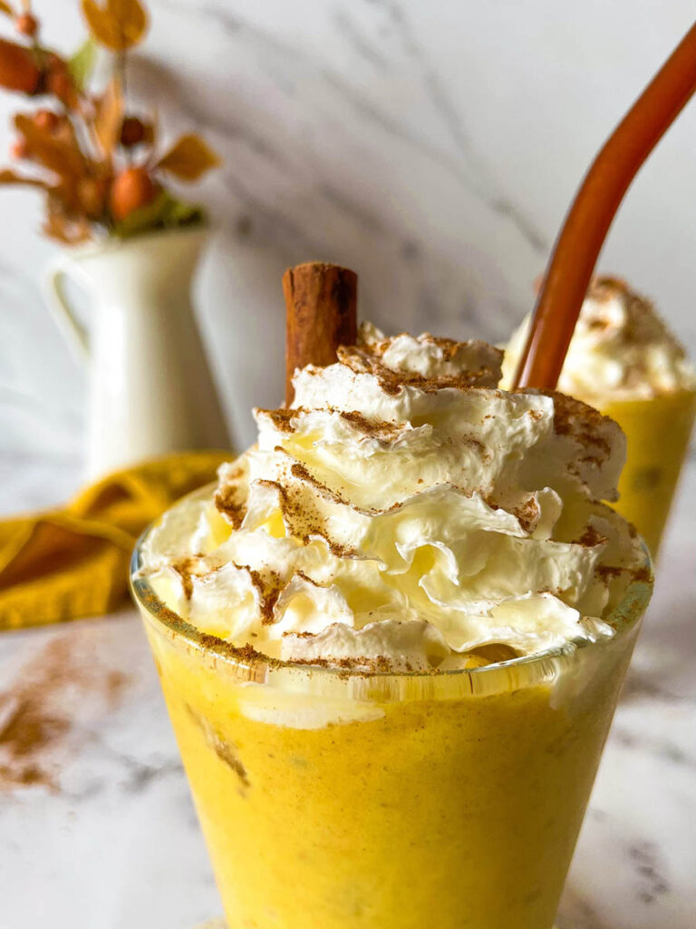 Irresistible Pumpkin Spice Mocktail Recipe 6 Close up detail of two tall glasses of creamy pumpkin spice mocktail topped with whipped cream and garnished with cinnamon sticks, set on a marble surface. A bundle of cinnamon sticks and a yellow cloth add cozy fall decor in the background, along with a small vase of autumn leaves.