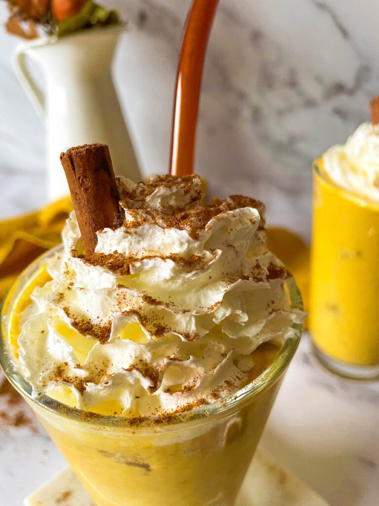 Irresistible Pumpkin Spice Mocktail Recipe 2 Close up detail of two tall glasses of creamy pumpkin spice mocktail topped with whipped cream and garnished with cinnamon sticks, set on a marble surface. A bundle of cinnamon sticks and a yellow cloth add cozy fall decor in the background, along with a small vase of autumn leaves.